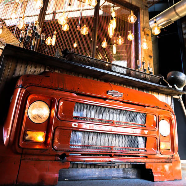 A vintage orange Chevrolet truck front, repurposed as decor, with Edison bulbs hanging above.