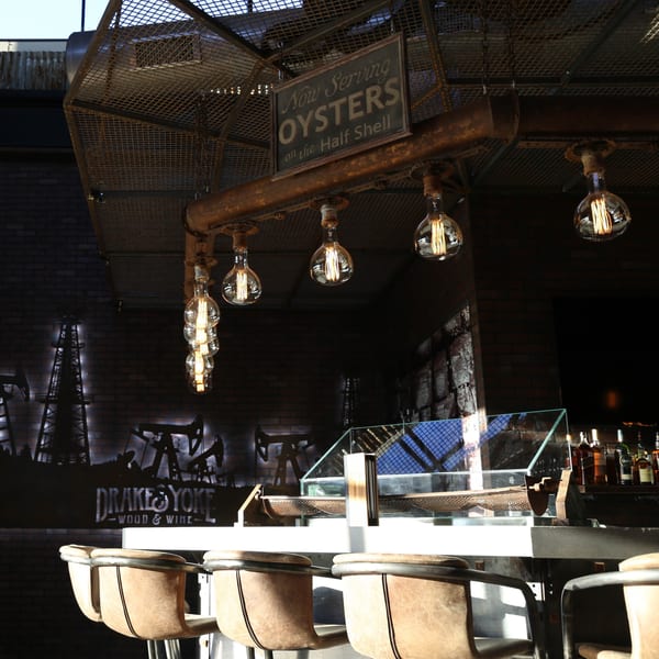 Drake's Yoke bar with industrial lighting, oyster sign, and oil rig mural on a brick wall.