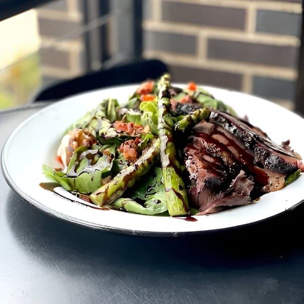 A white plate holds a BBQ steak salad with asparagus, greens, tomatoes, and balsamic glaze.